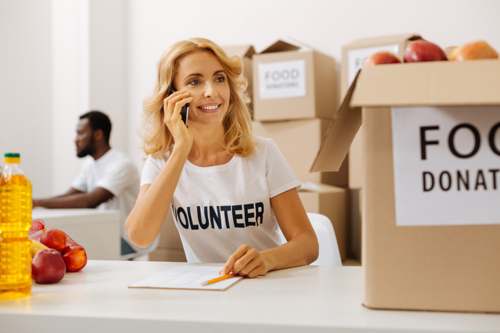 Woman volunteer talking to someone on the phone with donated food on the table and a man in the background using a computer.