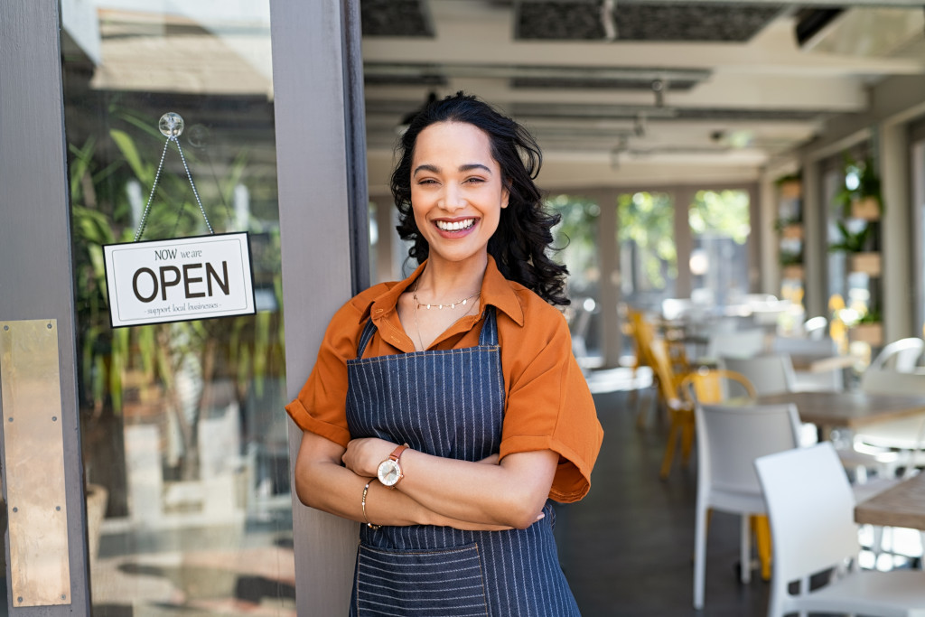 a woman in apron smiling and standing beside a sign that says open