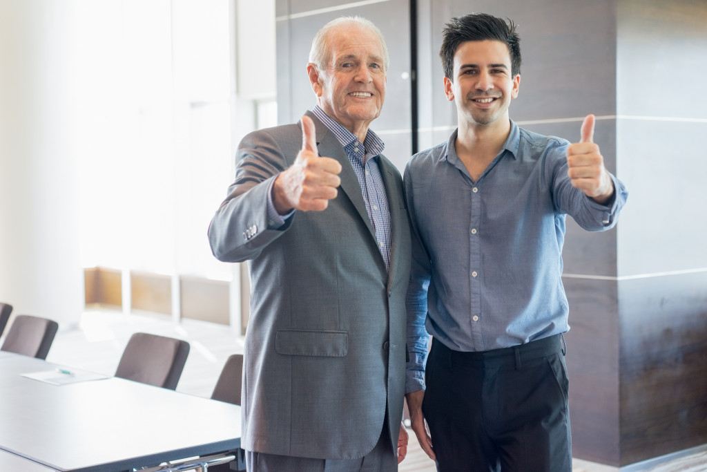 an old man and a young man both in business suits doing an okay sign and smiling