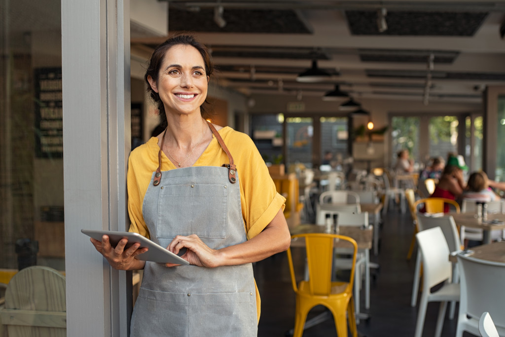 Smiling female business owner standing at the entrance of her restaurant holding a tablet.