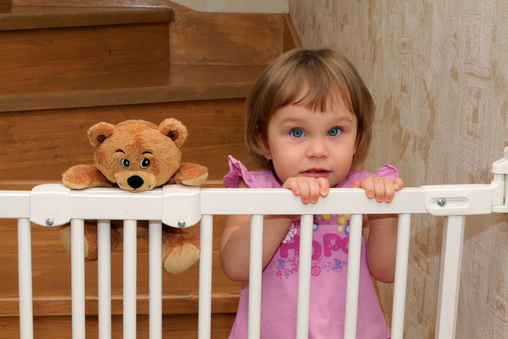 A young girl and her teddy bear behind a baby gate near the stairs